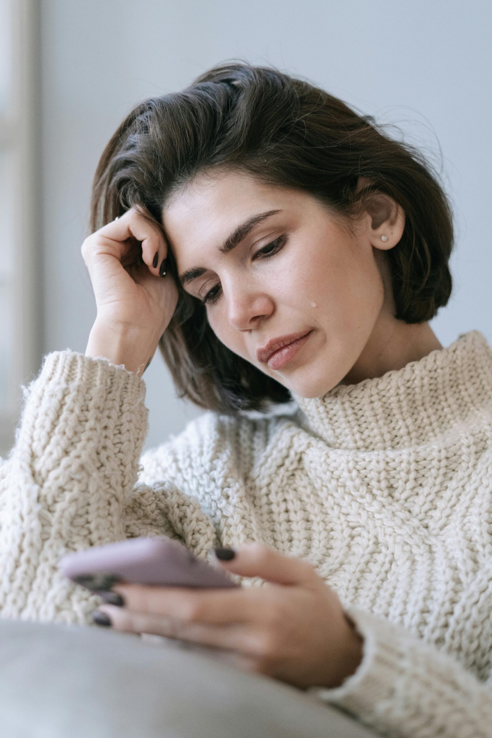A woman in a cozy sweater looks pensively at her smartphone, with a tear on her cheek, suggesting sadness or loneliness.
